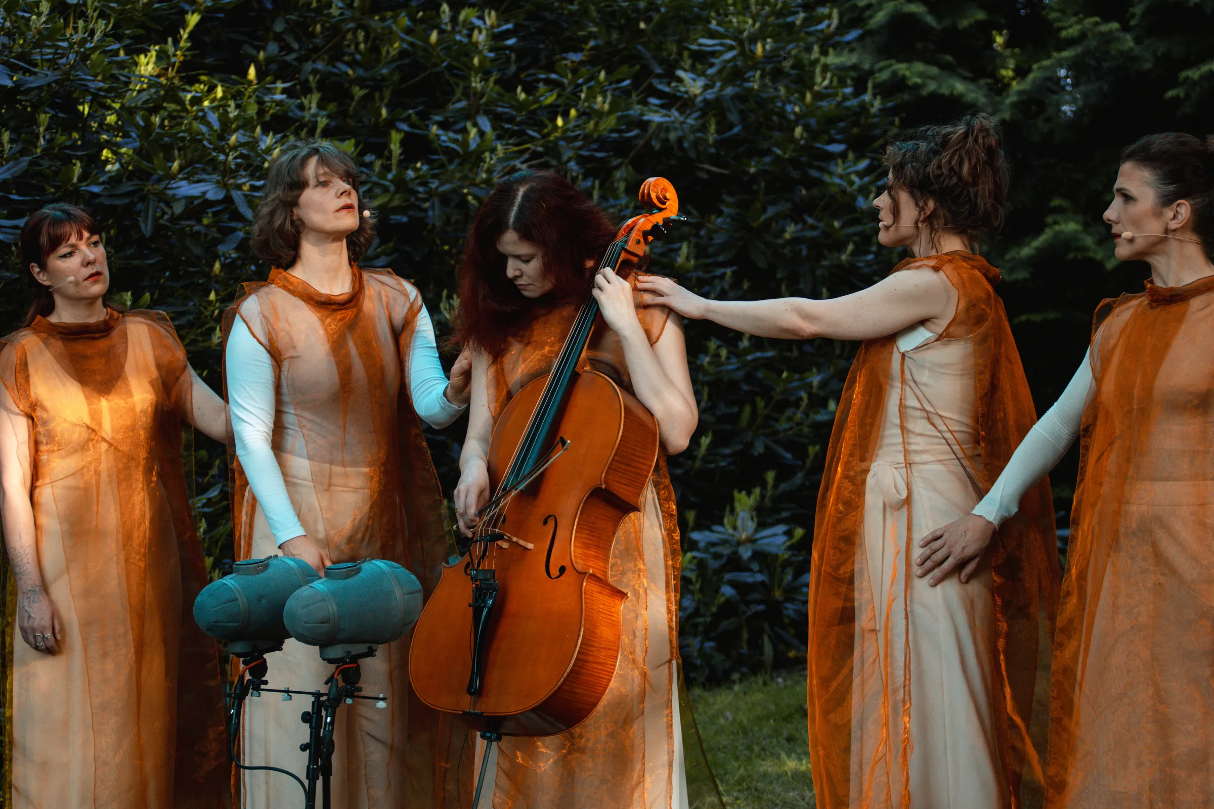 Maya stands with her eyes closed, holding a cello in her hands, surrounded by trees, with two singers on either side of her.