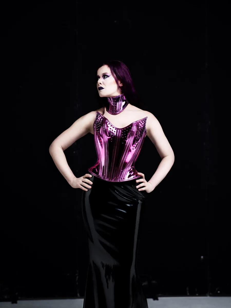 Maya Fridman standing hands on hips in a metallic pink corset and choker, black skirt, against a black backdrop