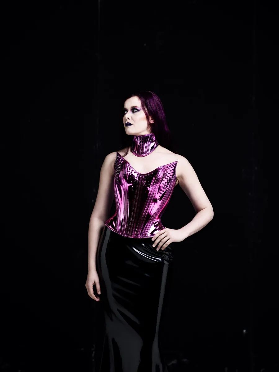 Maya Fridman in a metallic pink corset and choker, one hand on her hip, gaze lifted, against a black backdrop